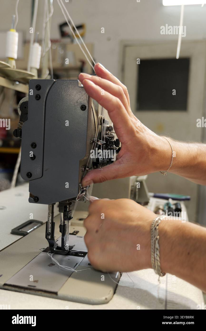 Hands carefully threading an industrial sewing machine, managing white thread and needles during the detailed process of preparing for production in a Stock Photo