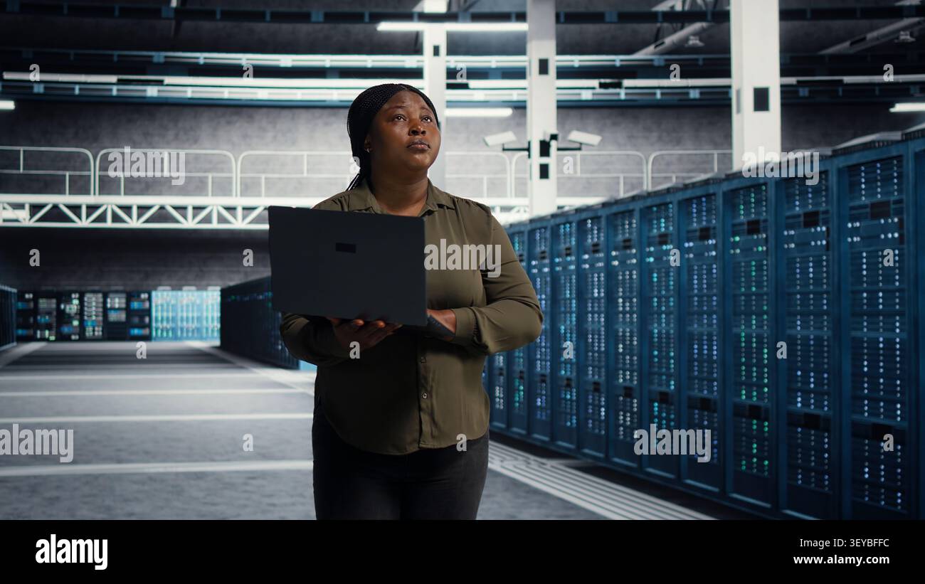 Cheerful systems engineer walking in server facility with notebook computer, checking hardware for system stability. Stock Photo