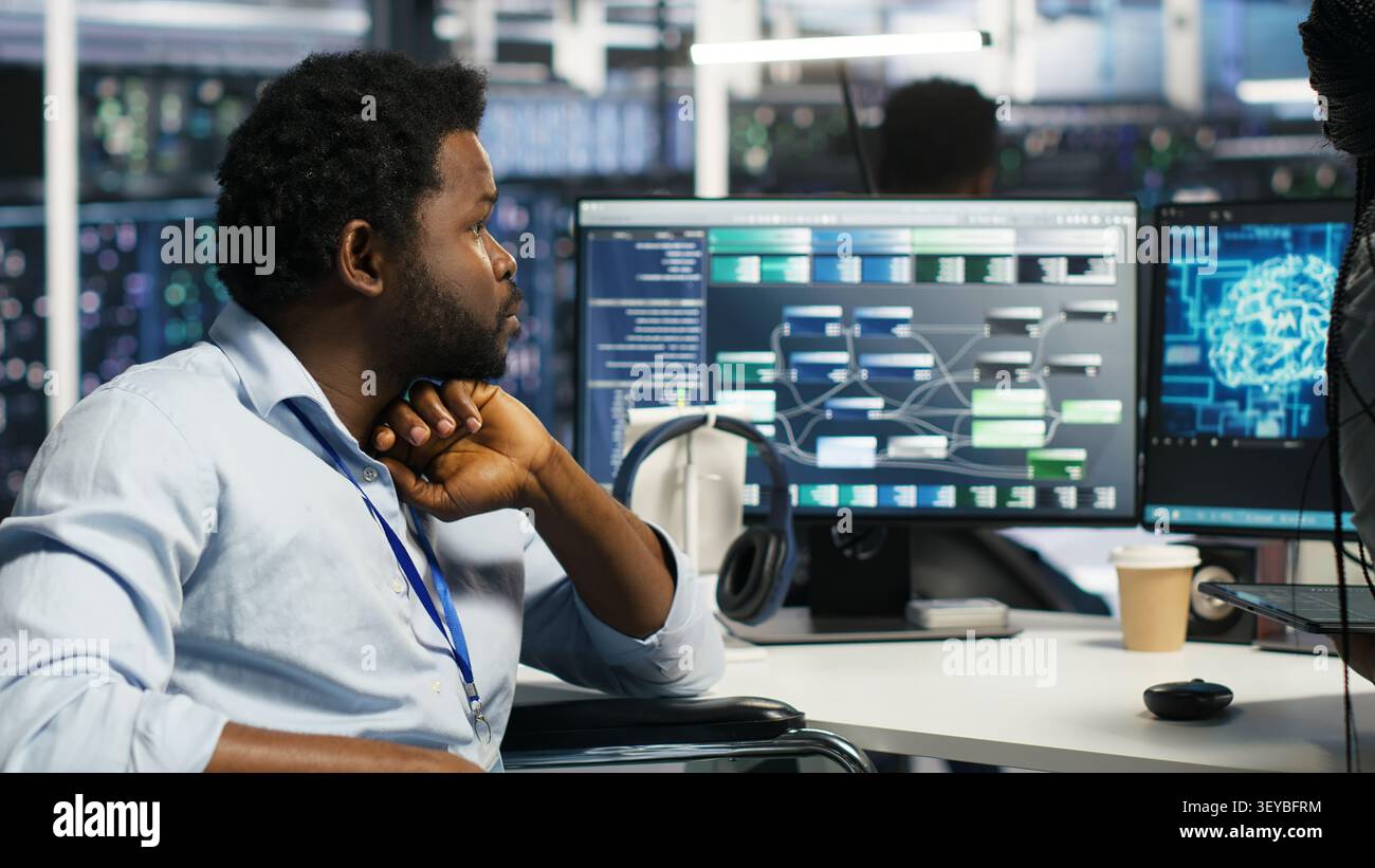 Server room engineer in wheelchair and colleague oversee automated tasks working with node tree programming tool. Stock Photo