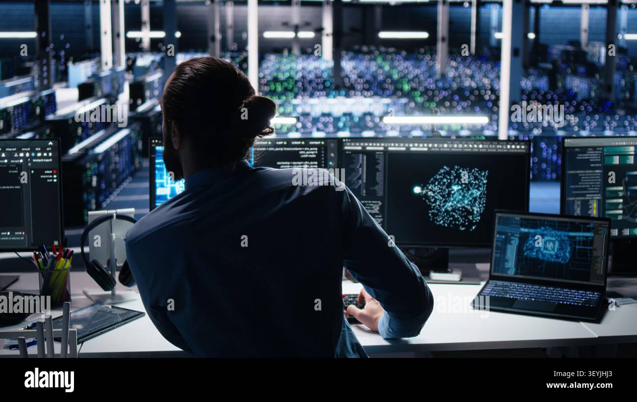 Computing facility computer scientist working with computer to track neural network working with AI large language model, while at work working with AI to oversee servers powering machine learning. Stock Photo