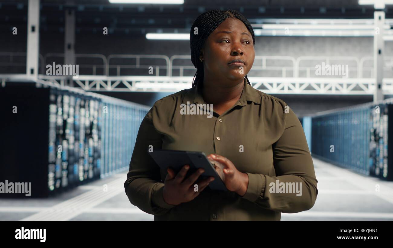 Admin walking in server facility, working with digital tablet to supervise software updates for system stability. Stock Photo