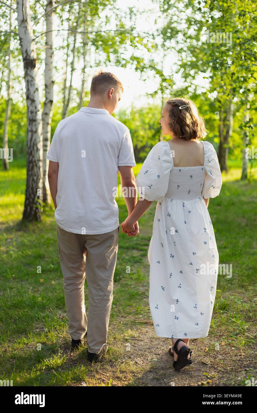 A young couple walks hand-in-hand on a dirt path surrounded by birch trees and green grass, bathed in warm sunlight Stock Photo