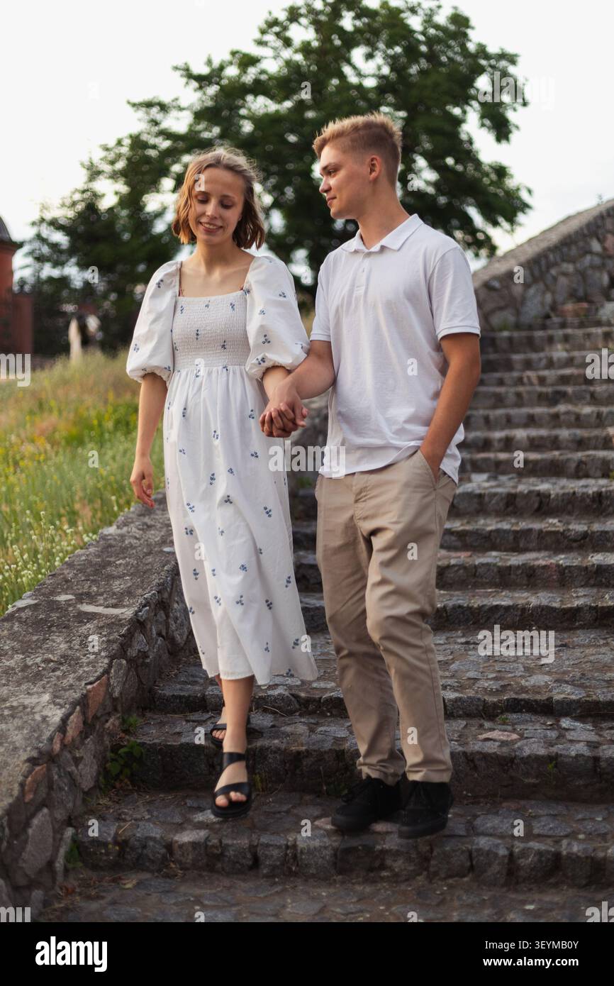 A teenage couple walks hand-in-hand down a worn stone staircase surrounded by greenery and an old stone wall Stock Photo