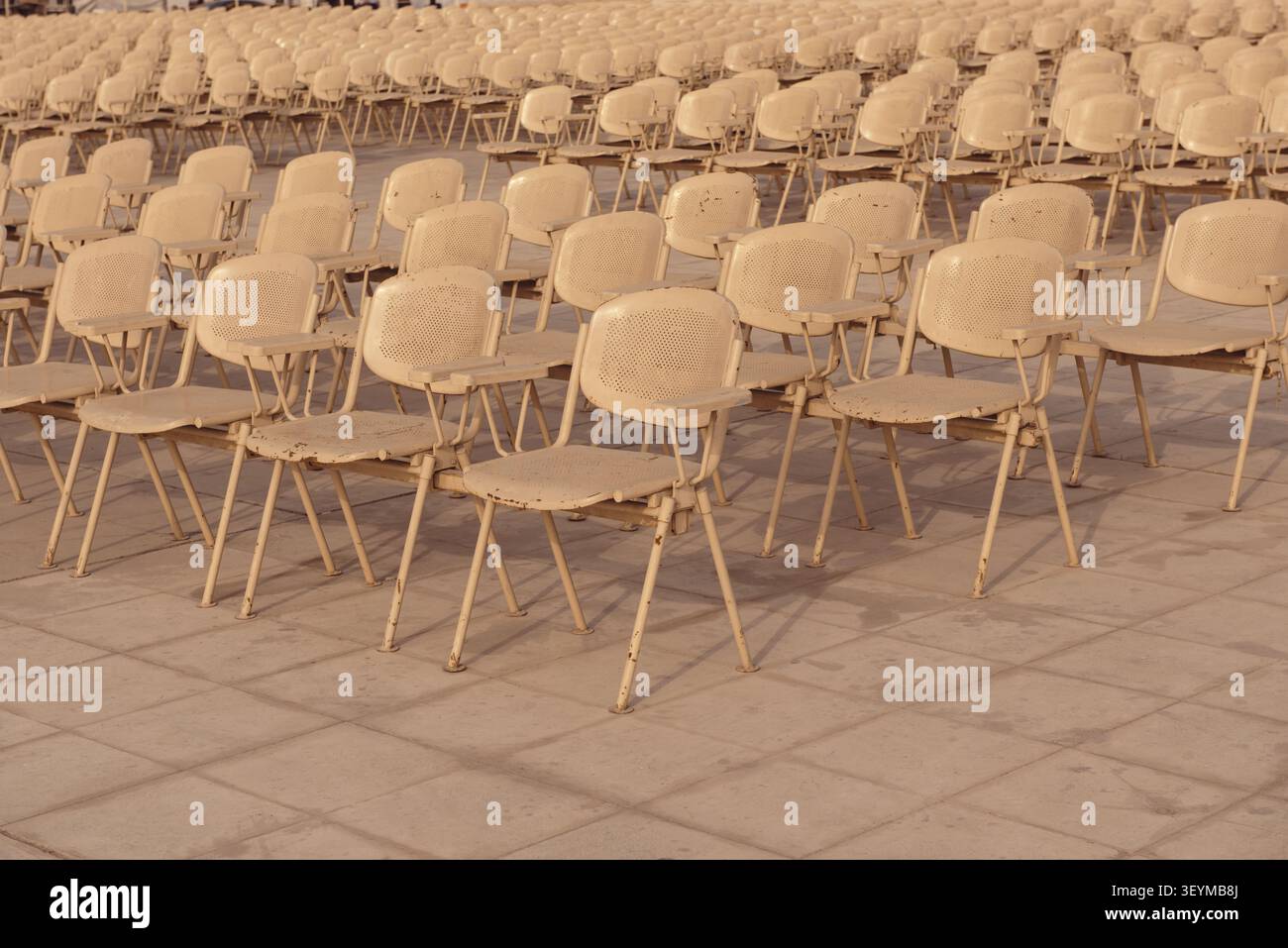 Vast rows of worn cream metal folding chairs arranged in a uniform grid on a stone plaza under warm golden hour light at an outdoor public event venue Stock Photo