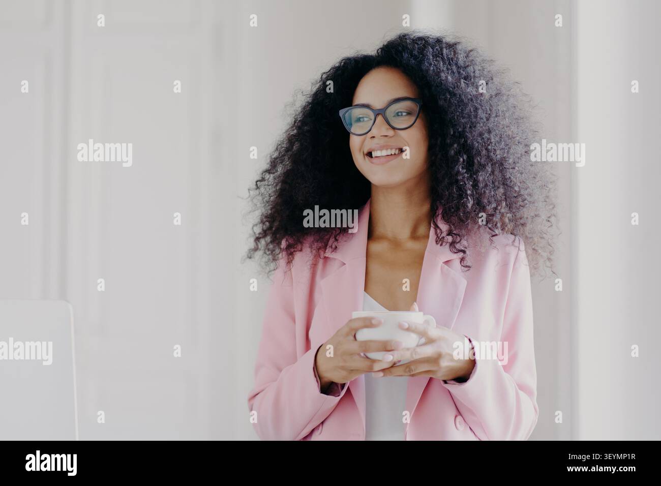 Photo of good looking African American prosperous busineswoman waits for partner in office, drinks coffee discusses future plans with colleague looks away with cheerful expression wears formal clothes Stock Photo