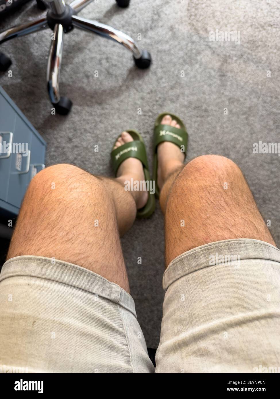 First-person perspective of a man wearing Hurley slides & denim shorts sitting indoors on a carpeted floor - Smartphone Captured Stock Image