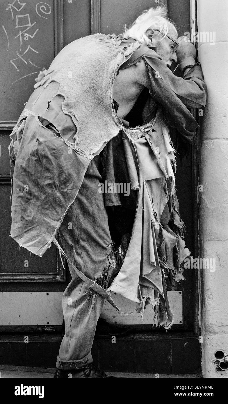 A black and white portrait of an elderly homeless man in tattered, heavily torn clothing standing hunched against a doorway in a city street. Stock Photo