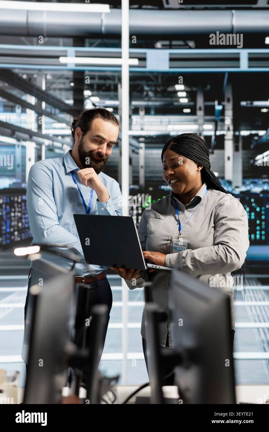 Cheerful team of engineers working with notebook computer in server environment, analyzing data. Stock Photo