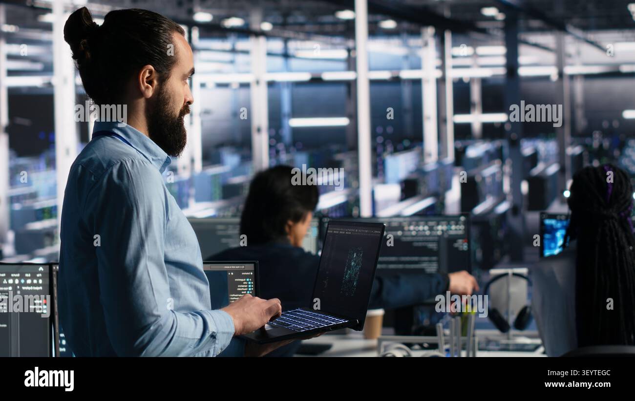 Data center technician monitors operational metrics on digital device inside a server room to improve infrastructure performance. Stock Photo