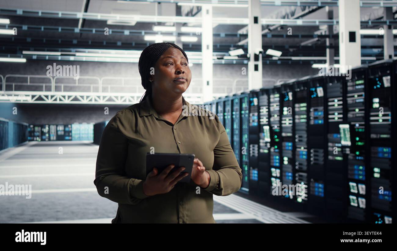 Admin walking in server facility, working with digital tablet to supervise software updates for system stability. Stock Photo