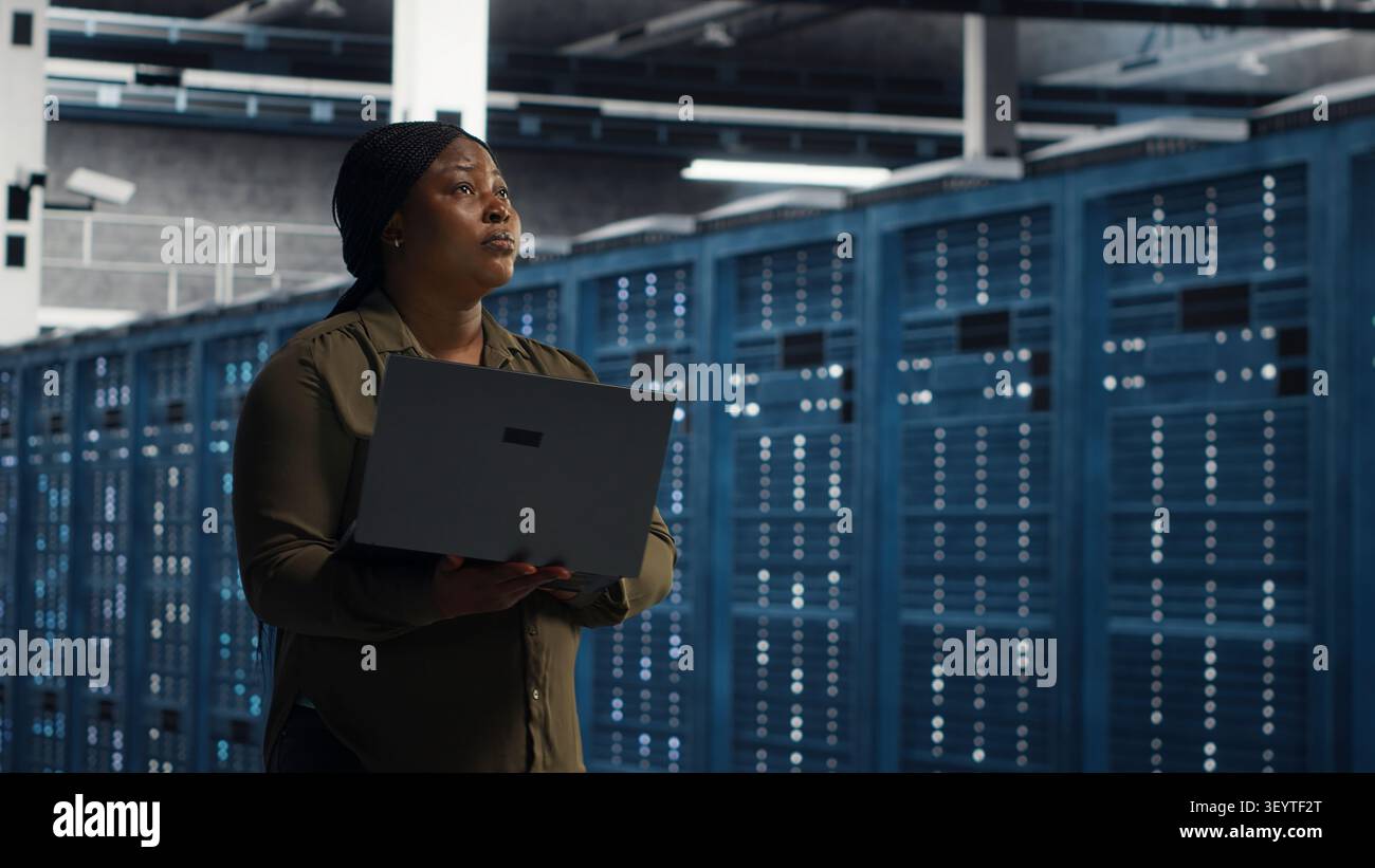 Cheerful systems engineer walking in server facility with notebook computer, checking hardware for system stability. Stock Photo