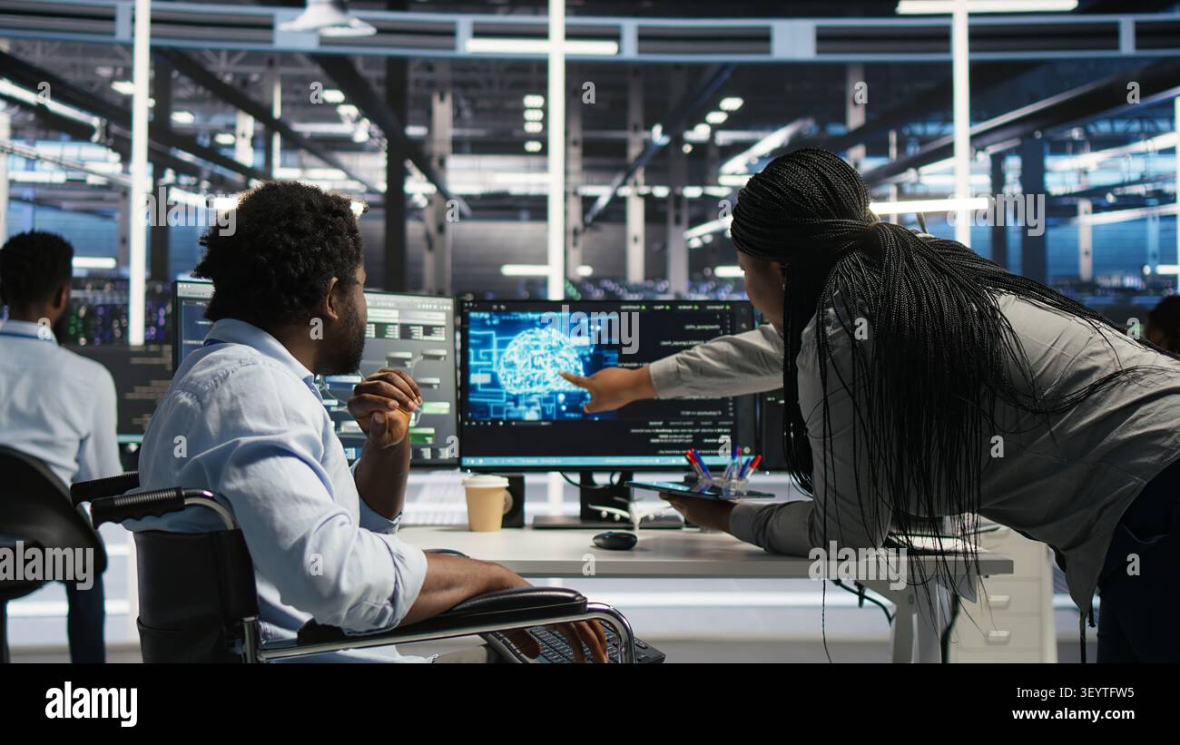 Server room engineer in wheelchair and colleague oversee automated tasks working with node tree programming tool. Stock Photo