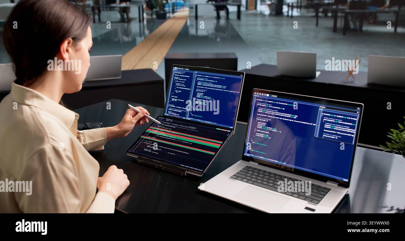 Young Female Software Developer Compares Code Changes On Dual Touchscreen Laptops. Stock Photo