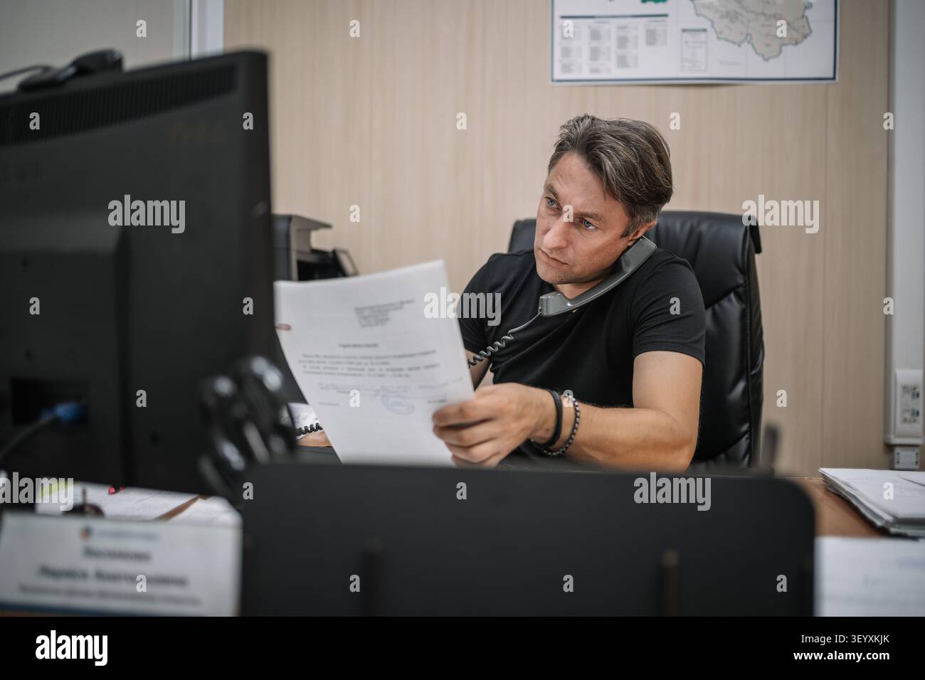 Office worker reading a document while talking on a landline phone. Multitasking, heavy workload, and focused business routine. Stock Photo