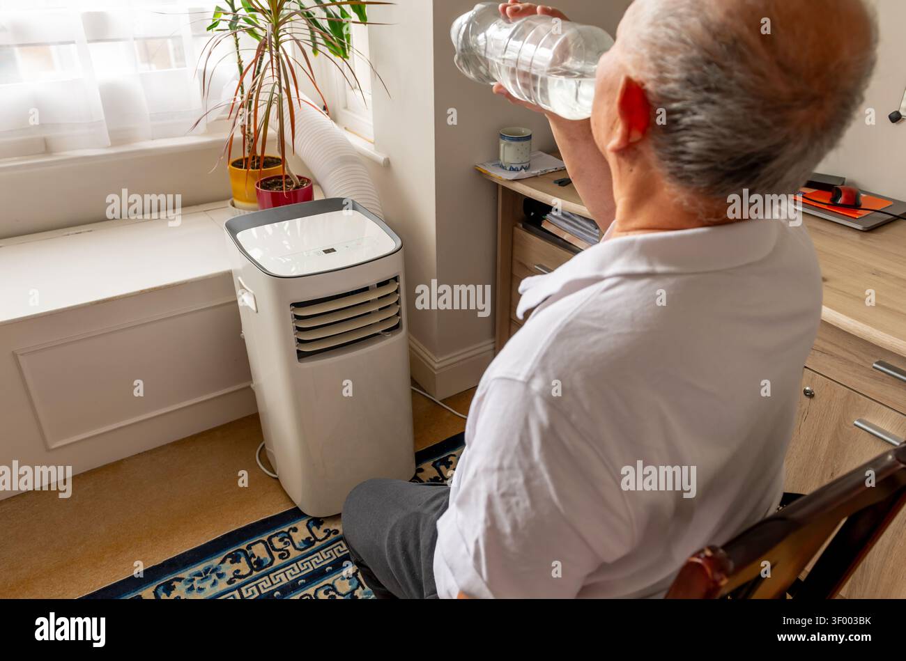 An elderly man cooling down in front of a portable air conditioner and drinking water during a Summer heat wave. Stock Photo