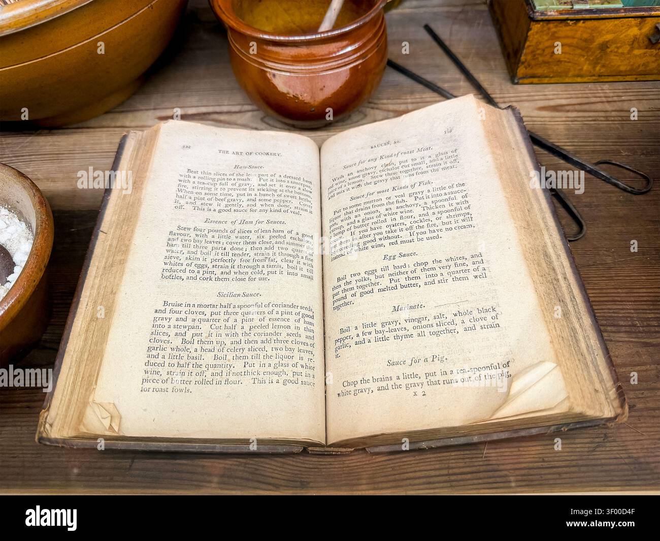 Open pages of The Art of Cookery, an old cookbook displayed on a wooden table with rustic pottery & kitchen utensils Stock Photo