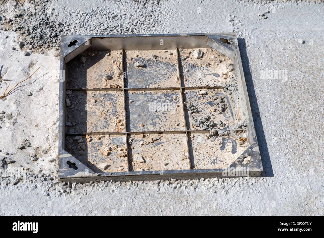 A close-up view of a square metal grate cover, partially filled with dirt and debris, set into a rough concrete surface at a building site Stock Photo