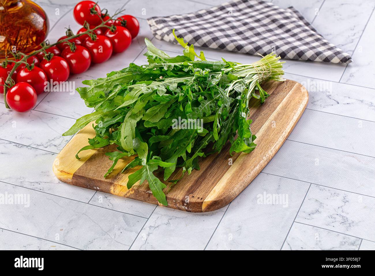 Fresh green arugula on wooden board beside red cherry tomatoes olive oil bottle. Gray marble countertop with checkered napkin. Natural light highlight Stock Photo