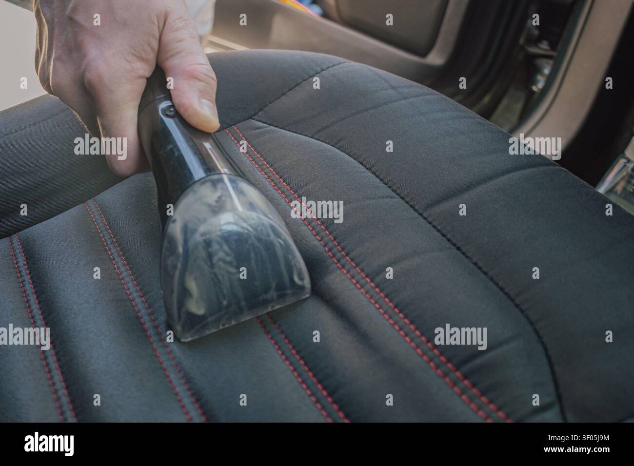 Horizontal close up shot of a man cleaning a black fabric car seat upholstery with red stitching details using a professional vacuum water extraction Stock Photo