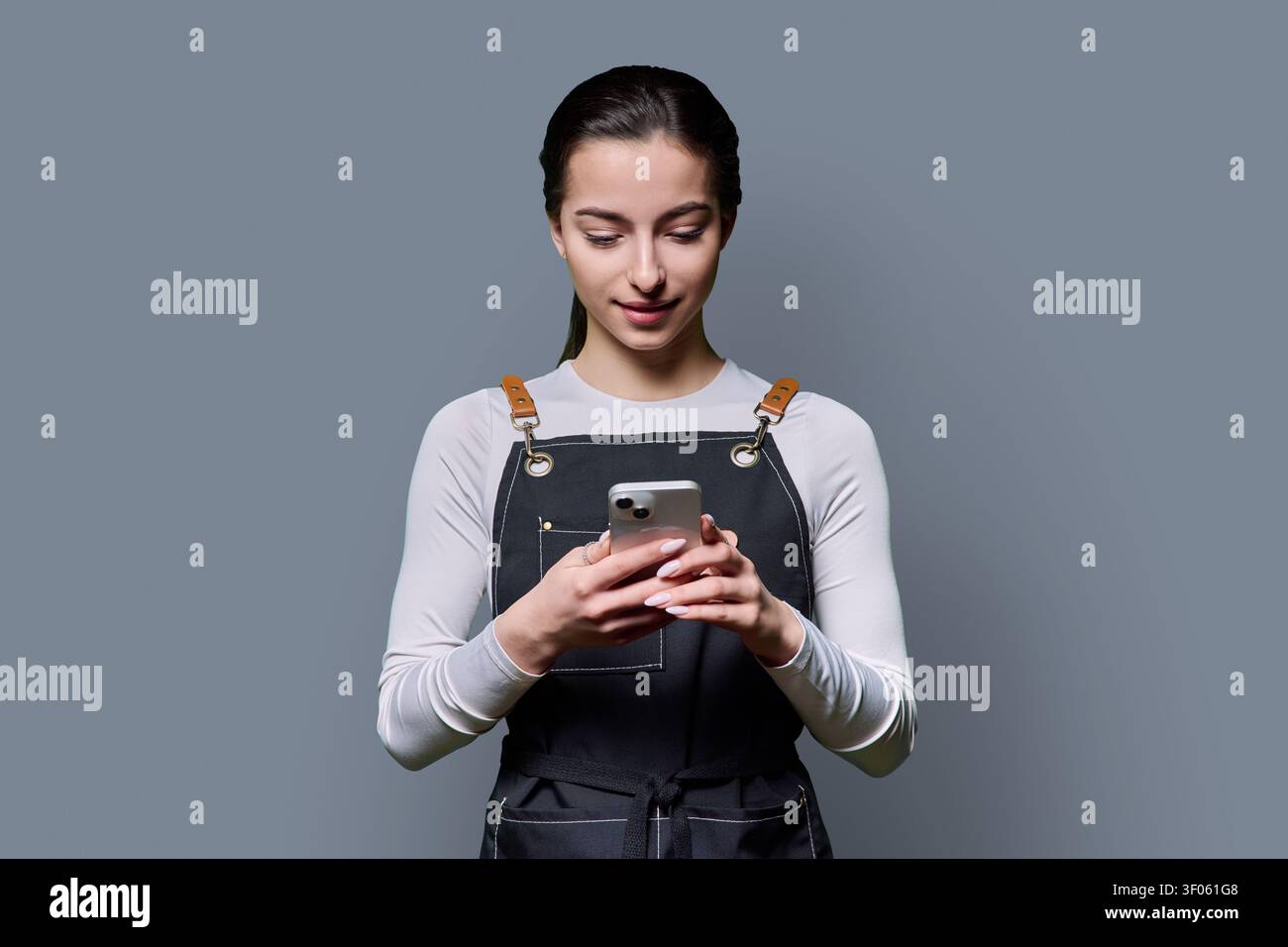 Young female in apron looking down at smartphone screen on gray background Stock Photo