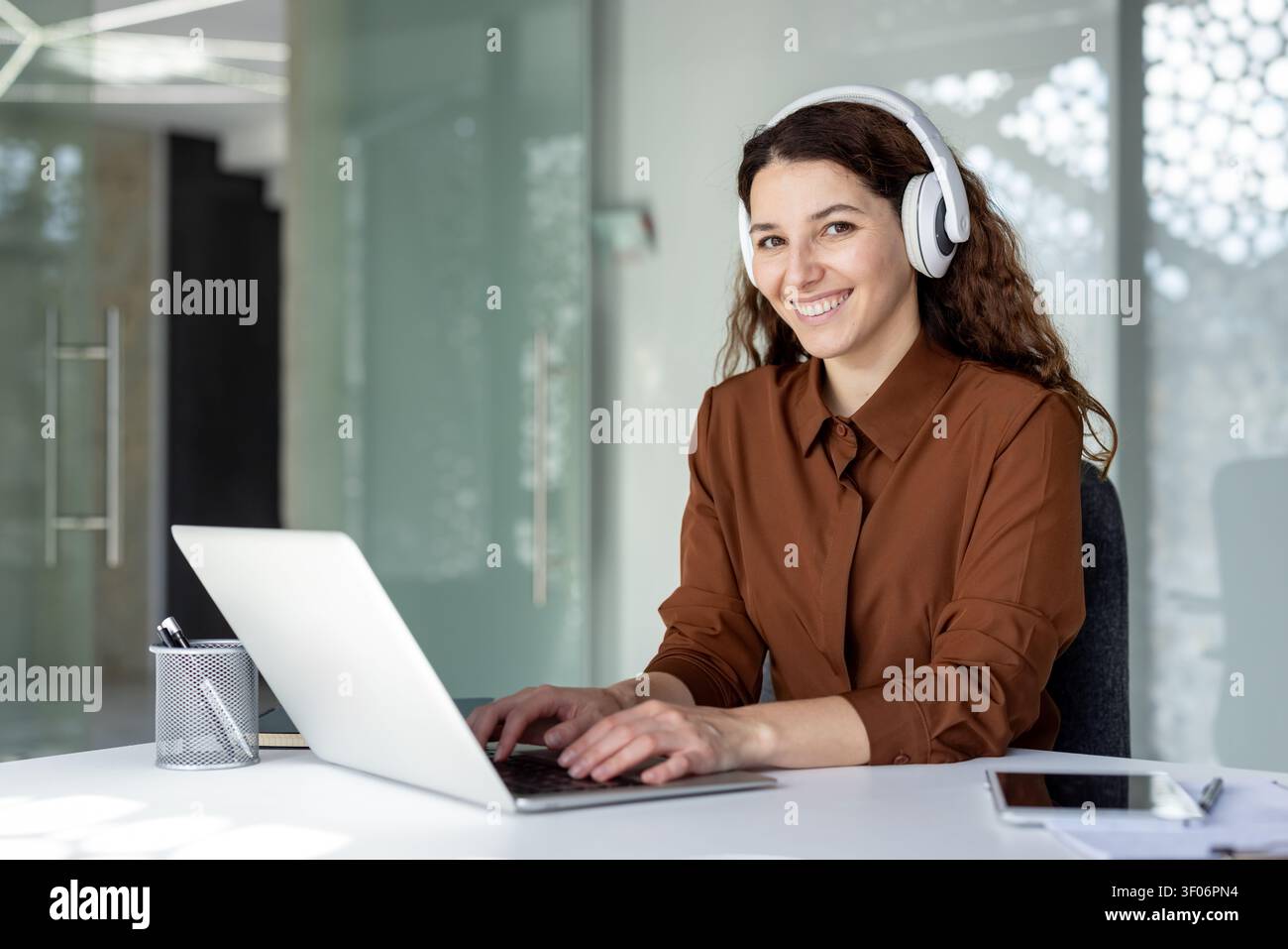 Young businesswoman smiling while typing on her laptop at an office desk, wearing white headphones and providing remote customer support Stock Photo