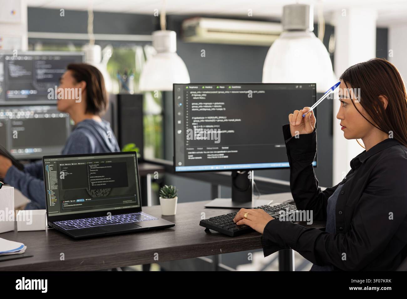 Female software developer taps pen on forehead, studying coding scripts on screen with thoughtful expression. Diligent asian woman works on computer, brainstorming ideas for app development. Stock Photo