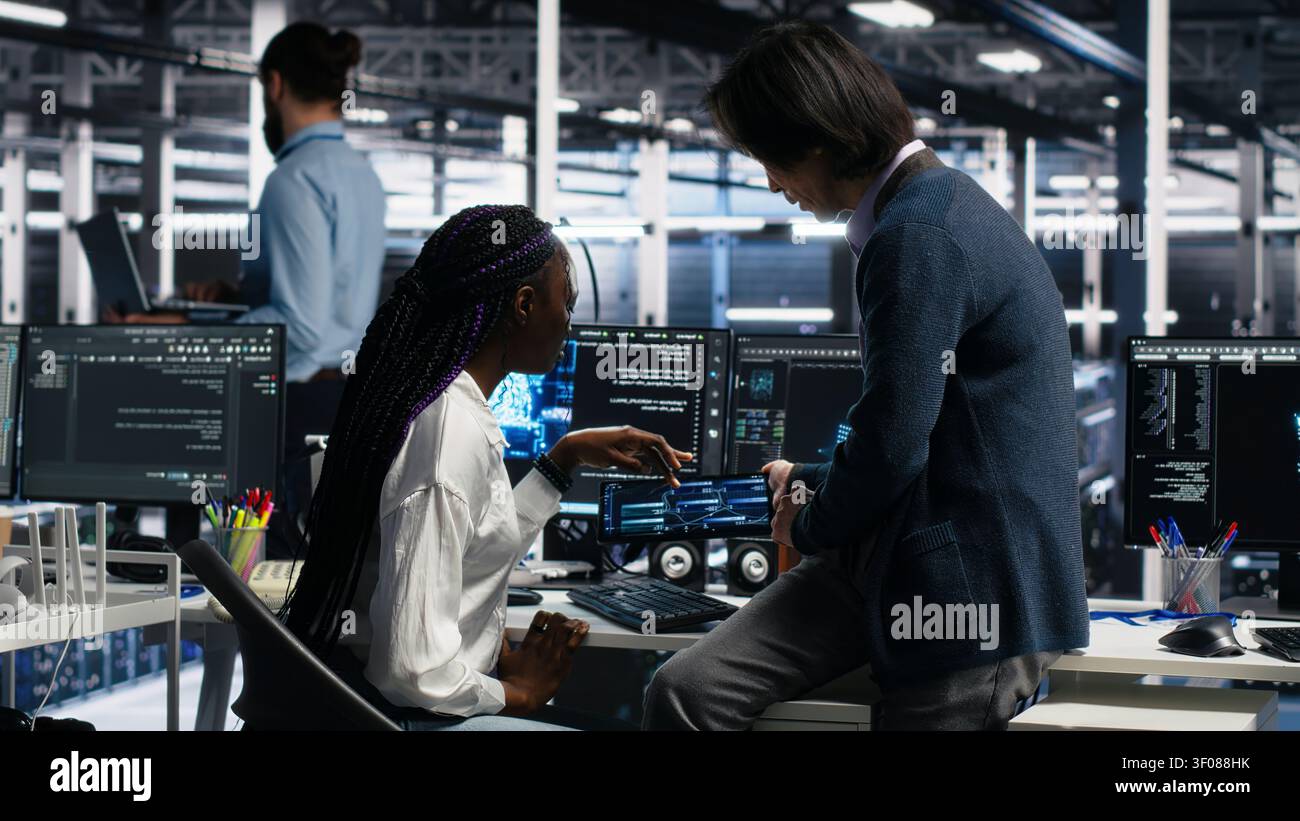 Lead engineer and trainee use a PC in a server room to develop neural network software and implement machine learning technology. Stock Photo