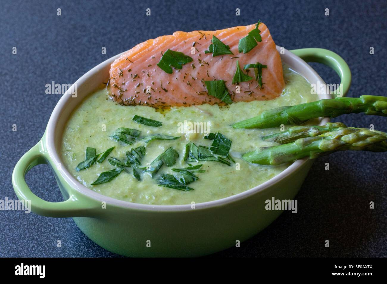 Creamy asparagus soup topped with salmon fillet, dill and parsley in a green casserole, asparagus tips on the right, close angle, dark surface Stock Photo