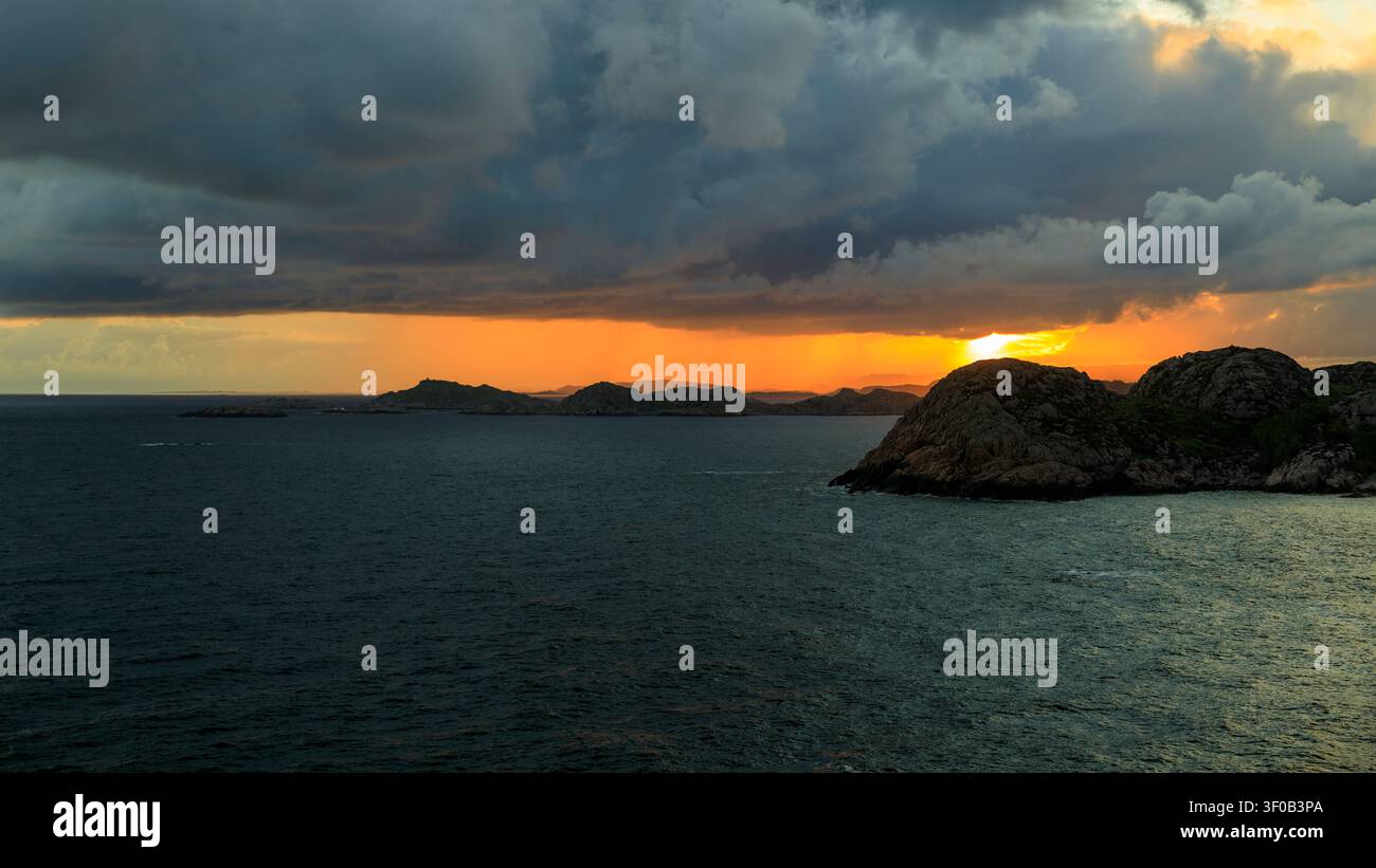 Golden sunlight pierces through heavy dark storm clouds over rocky islets in the sea at sunset in Lindesnes, Norway Stock Photo