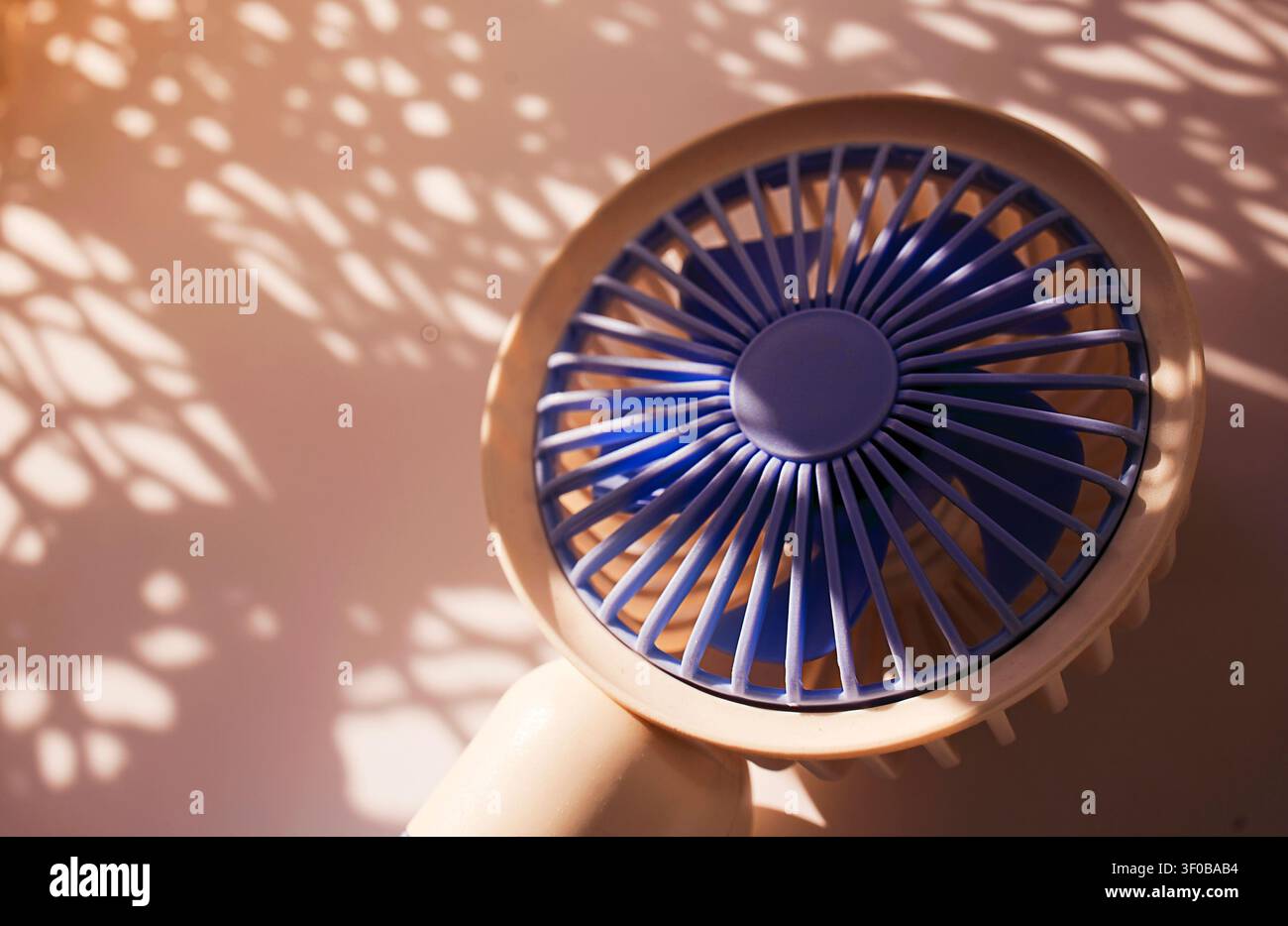 Handheld fan in blue and beige colors creates detailed shadows on a light surface, highlighting its design and structure in a well-lit setting Stock Photo
