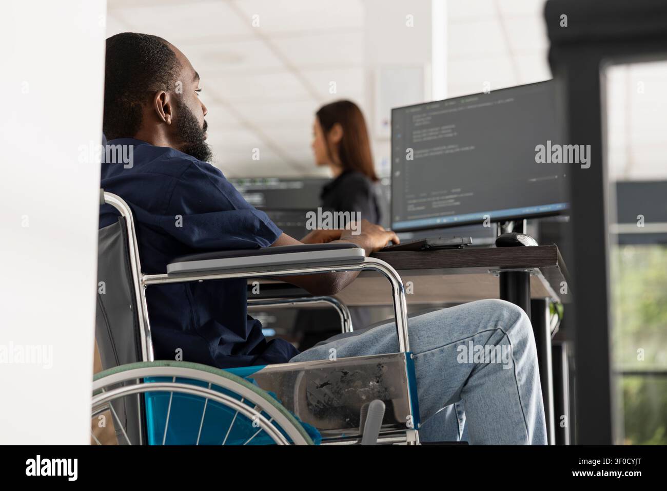 Male wheelchair user works at desktop PC, debugging software and reviewing website code. Dedicated black man developer completes programming tasks in inclusive tech startup environment. Stock Photo
