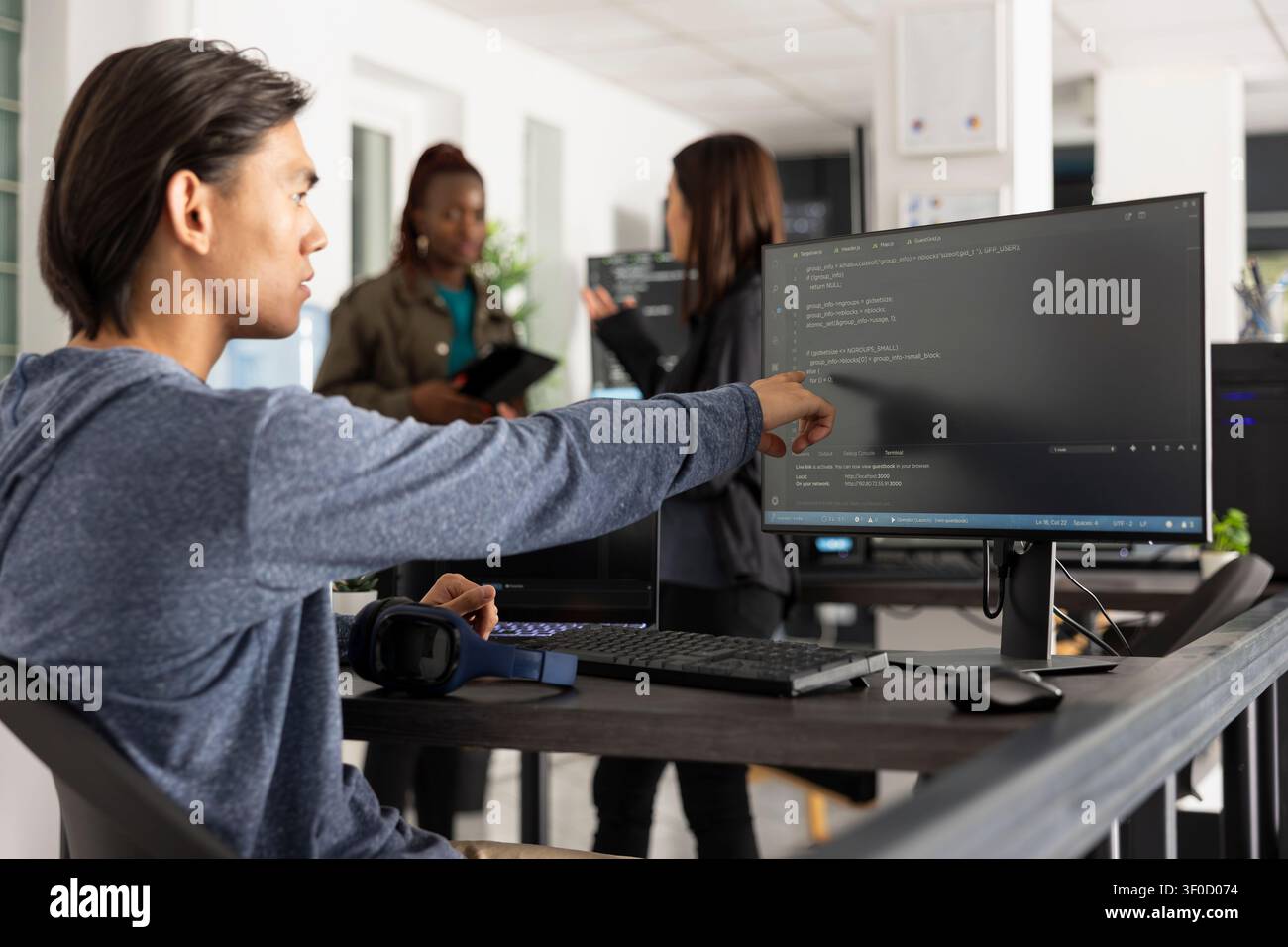 Asian male programmer points at desktop monitor with surprised expression after identifying key code statement to solve web development issue. Man reviews HTML scripts in modern tech workspace. Stock Photo