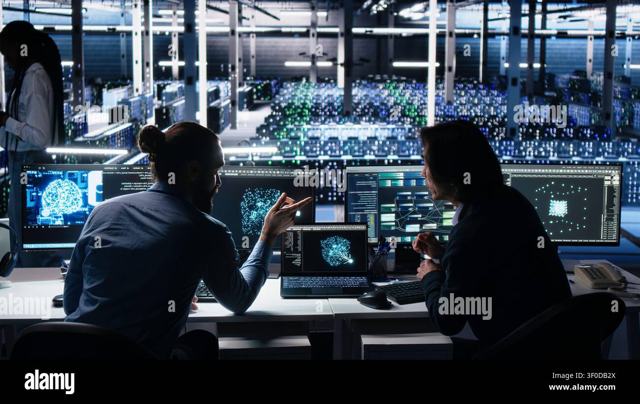 Senior programmer and coworker programs on a computer workstation in a data center with generative AI assistance. Stock Photo