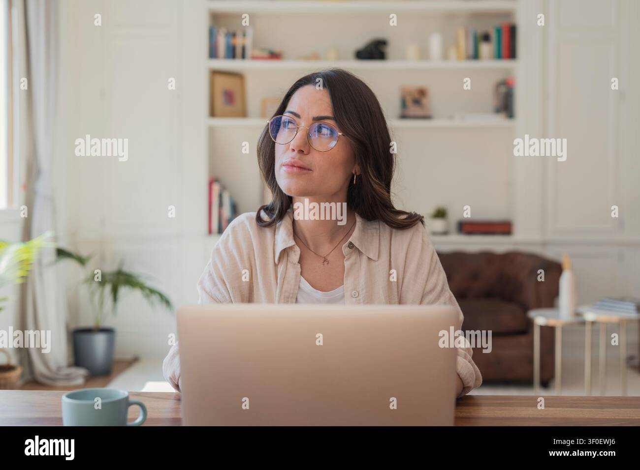 A thoughtful woman wearing glasses works on a laptop in a bright home office, pausing to look up as she thinks. Cozy workspace with bookshelf, sofa, c Stock Photo