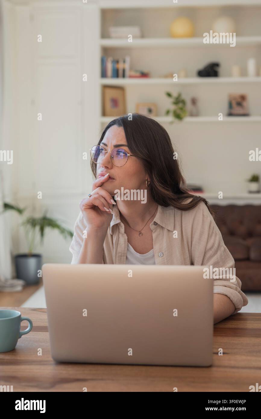 Young woman wearing glasses works on a laptop in a tidy home office, looking thoughtful as she plans and researches remote projects. Natural light, co Stock Photo