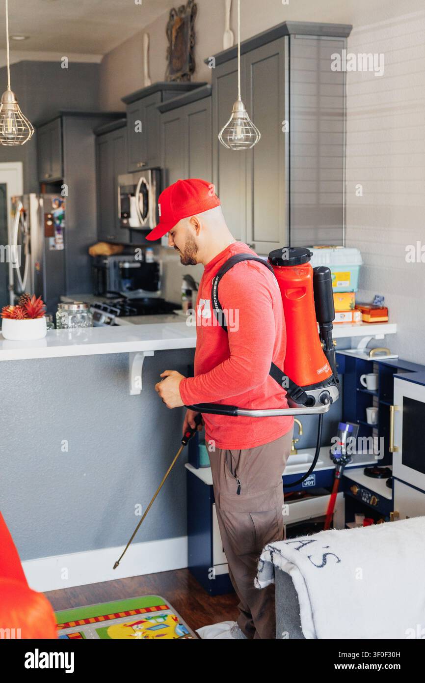 Man in red cap sprays kitchen with professional equipment. He wears backpack sprayer and focused expression. Bright lighting shows clean, modern kitch Stock Photo