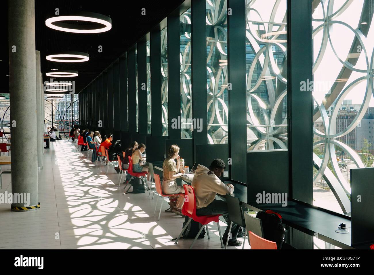 Students studying by large windows with patterned shadows inside the Library of Birmingham, UK Stock Photo