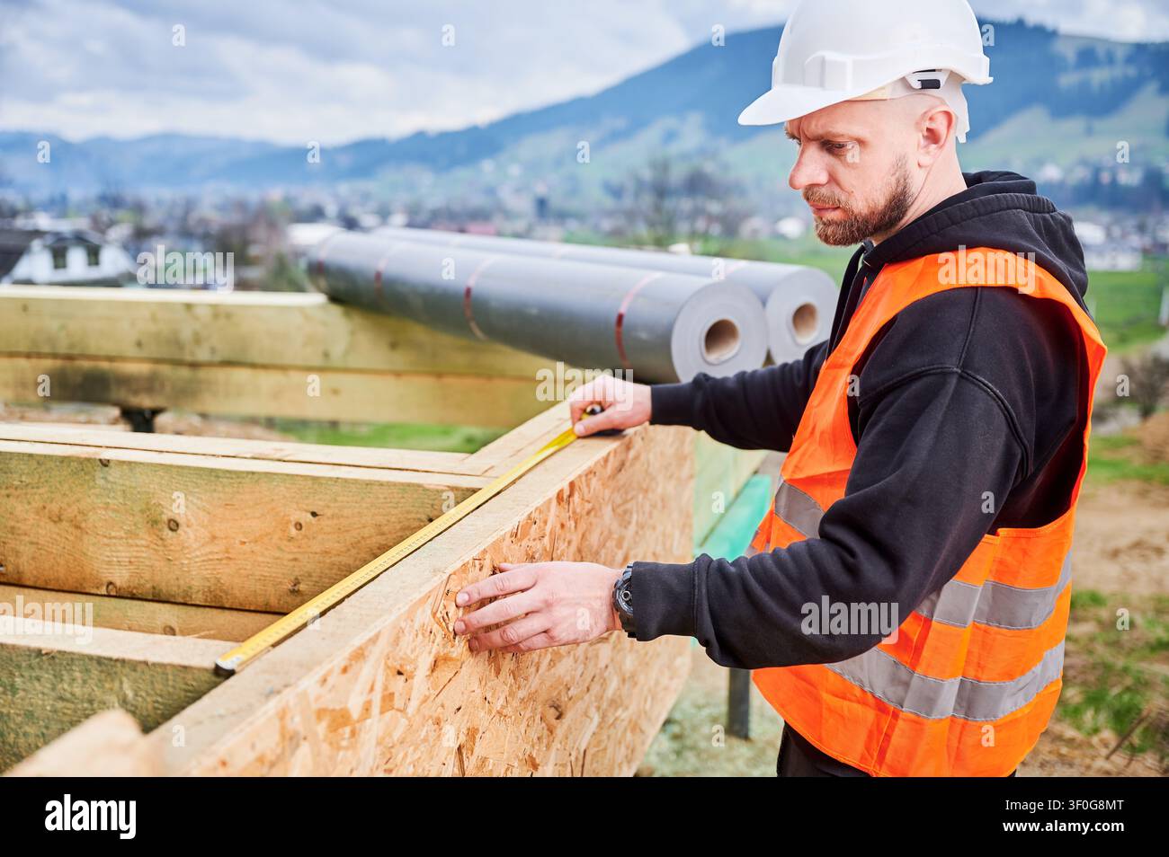 Construction worker in safety vest measuring wooden wall panel with tape, checking dimensions and alignment during house building process on outdoor site Stock Photo