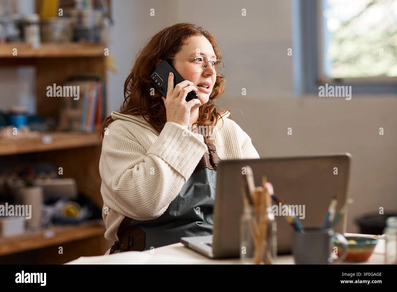Pottery Studio Small Business Owner Talking on Phone While Working on Laptop in Creative Workshop Stock Photo