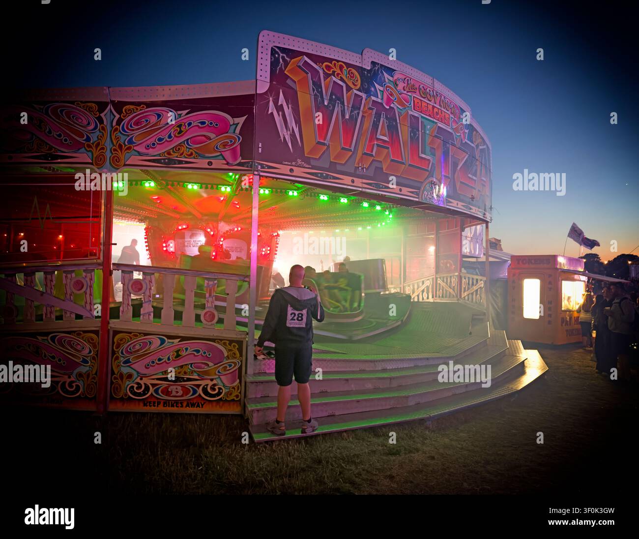 Illuminated Waltzer fairground ride at dusk, with a waiting participant beneath the familiar slogan “scream if you want to go faster” Stock Photo