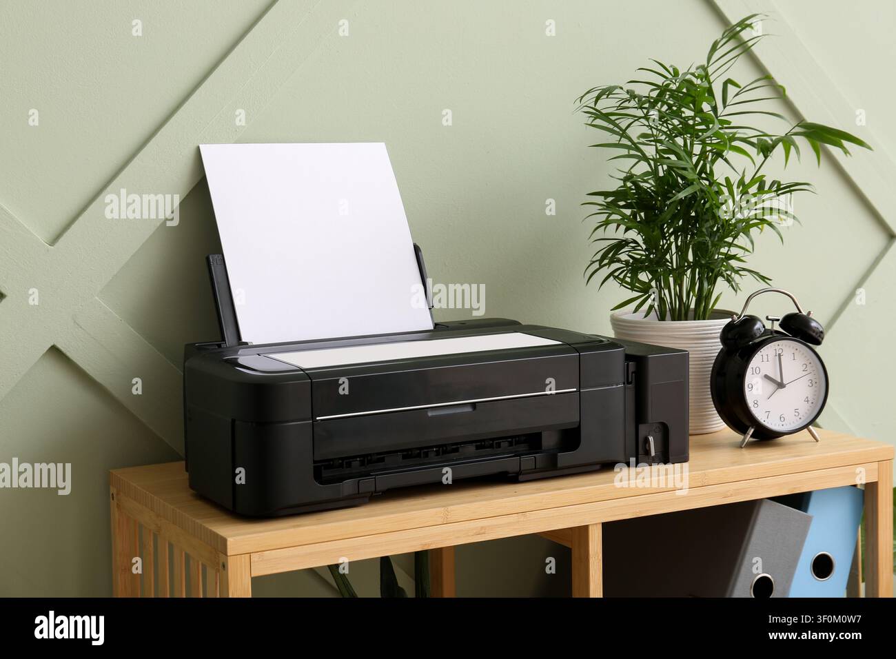 Black inkjet printer with blank paper on a wooden shelf next to a potted plant and alarm clock, the background features a sage green wall with a subtl Stock Photo