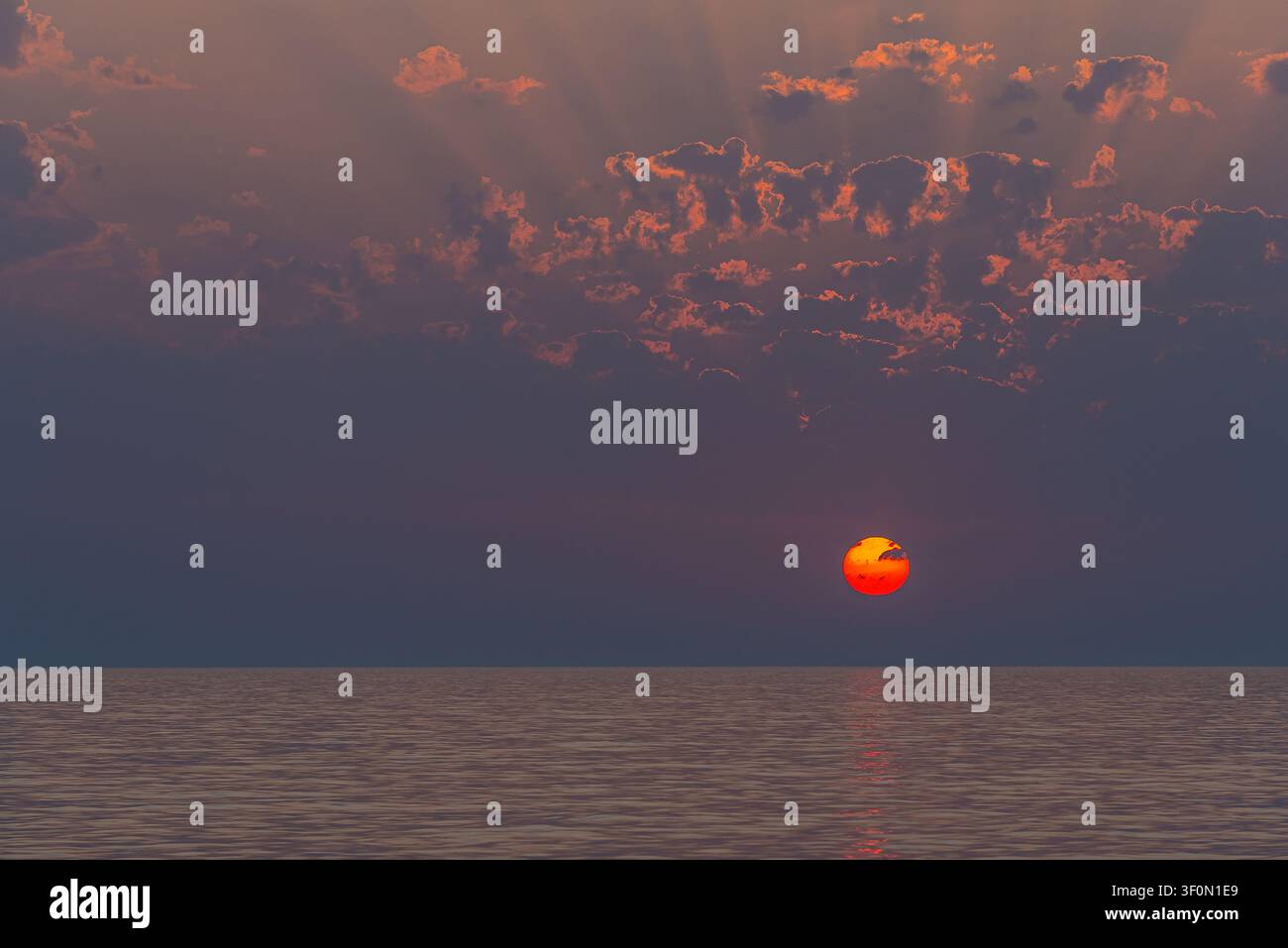 A vivid red solar disk hangs just above the horizon over a calm, glassy sea. Stock Photo