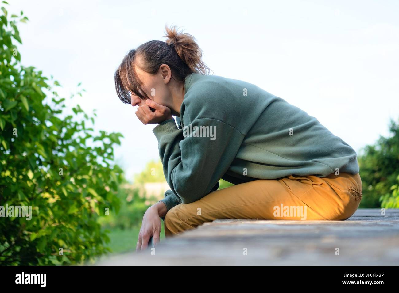 A young woman sits on a bench with her head in her hands in a green garden. The woman hiding her face presents a lonely, despair moment of emotional distress and thoughtful reflection outdoors. Stock Photo