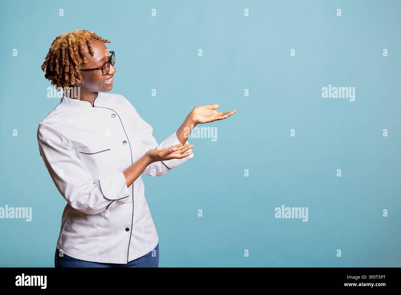 Cheerful female chef gesturing toward empty space against blue background. Professional cook with open hands, presenting mockup area for restaurant promotions, branding and culinary marketing visuals. Stock Photo