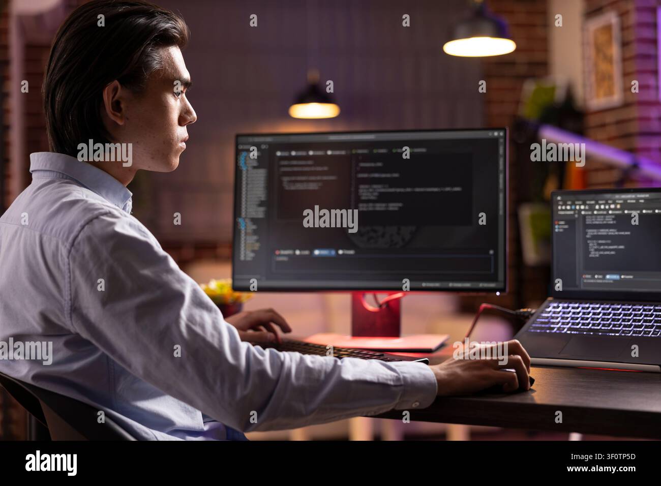Young male freelancer sits in home office during evening hours, analyzing and debugging code on laptop and desktop PC. Asian entrepreneur developing web applications for online company. Stock Photo