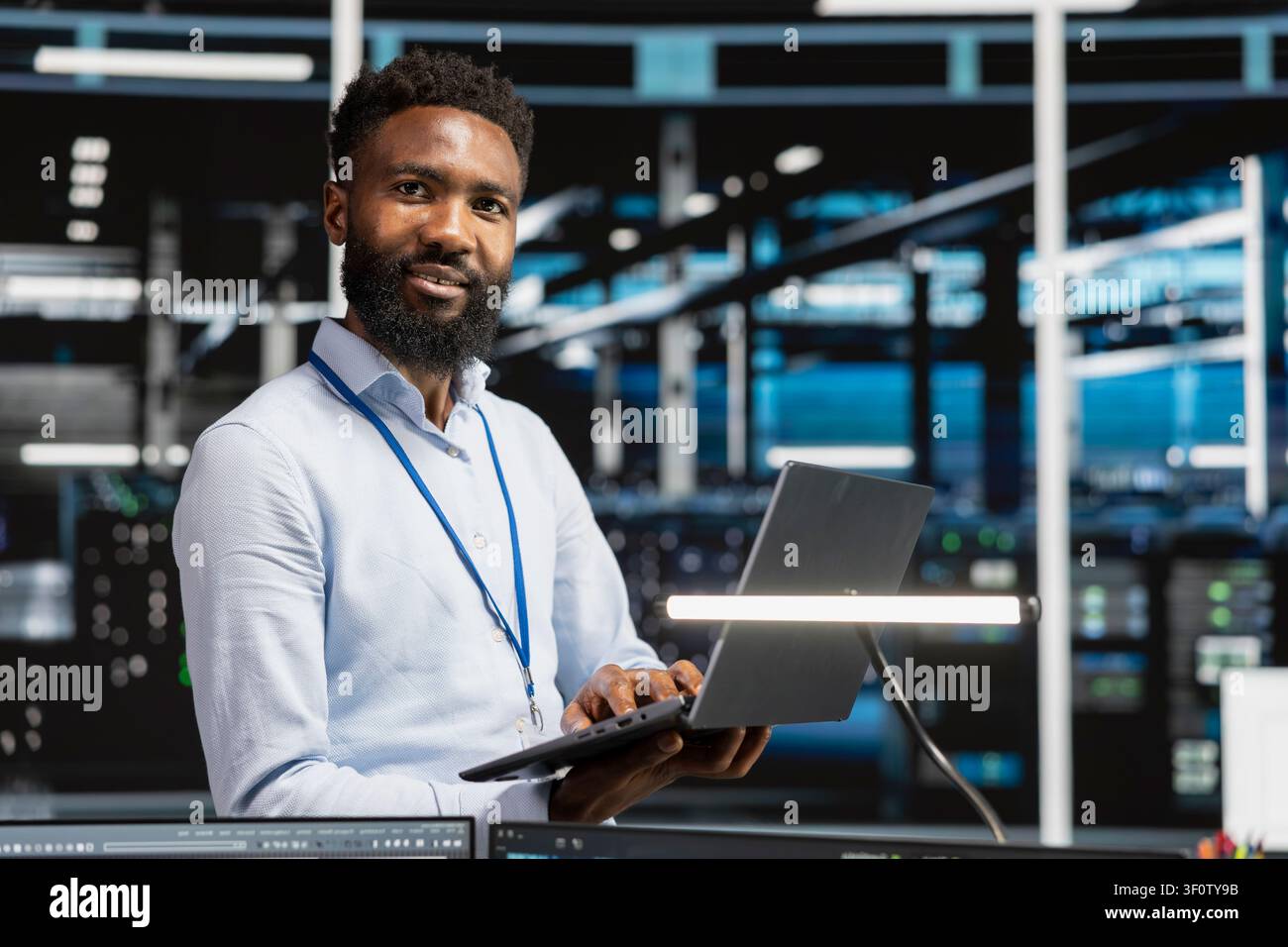Cheerful data center engineer monitors system performance on a laptop, identifying bottlenecks and troubleshooting server hub errors. Stock Photo