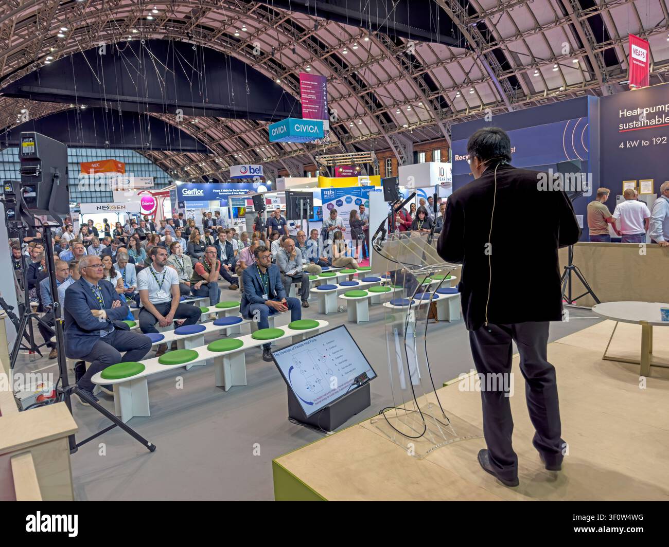 Speaker addresses a seated audience on a Housing 2023 session stage at Manchester Central, with housing-sector exhibition stands behind Stock Photo