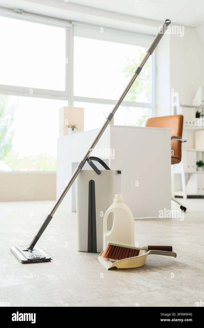 Cleaning supplies including a mop, bucket, detergent, brush, and dustpan on a light floor in an office setting Stock Photo