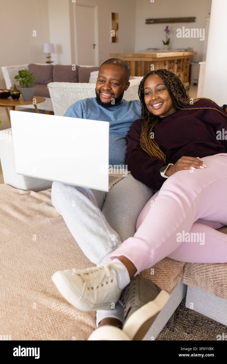 African couple sitting on light-colored sectional sofa, leaning close and viewing white laptop Stock Photo
