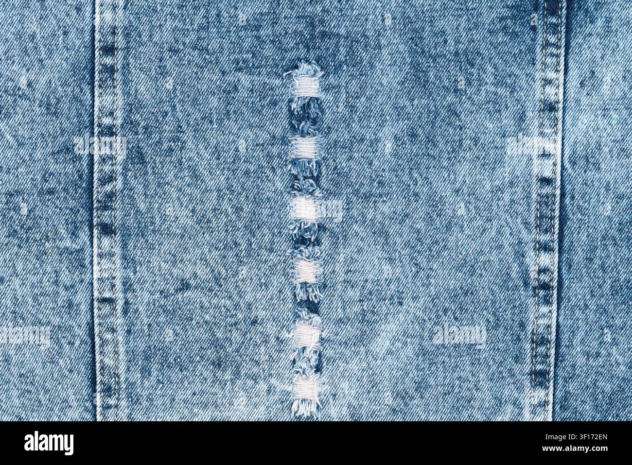 Light blue denim texture background. Distressed fabric with vertical rip, frayed white threads, and double seams. Casual, rugged clothing material for Stock Photo